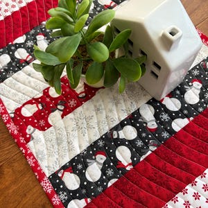 May include: A quilted table runner featuring alternating diagonal stripes of red, white, and black fabric. The pattern includes printed motifs of snowmen wearing hats and scarves, along with white snowflakes on red and black backgrounds.