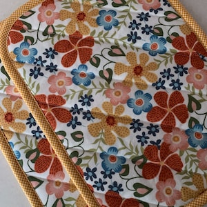 May include: Two quilted pot holders with a floral pattern. The pot holders have a white background with red, yellow, blue, and pink flowers. The edges of the pot holders are trimmed with a yellow and white checkered fabric.