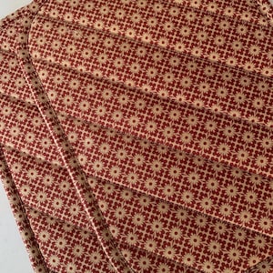 May include: A quilted fabric potholder featuring a repeating pattern of small cream-colored flowers on a deep red background. The item has diagonal stitched lines across the surface and a rounded edge with matching fabric binding along the border.