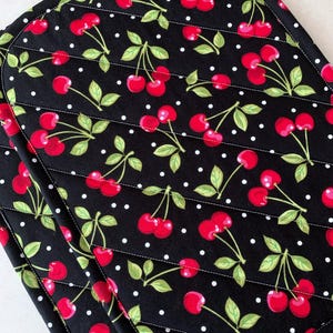 May include: Black quilted placemats with a repeating pattern of red cherries and green leaves on a black background with white polka dots. The placemats have a rounded shape and white stitching.