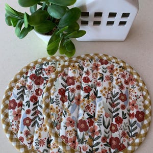 May include: Two round floral print pot holders with a yellow and white gingham trim. The pot holders are quilted and have a white background with red, pink, and yellow flowers.