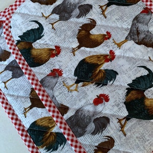 May include: Two quilted pot holders with a rooster print. The fabric features roosters in various colors, including brown, gray, and gold, set against a white background with a subtle newspaper-like pattern. The edges are trimmed with a red and white gingham fabric.
