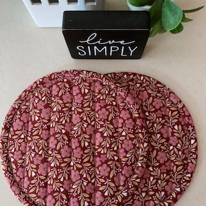 May include: Two round, quilted fabric pot holders featuring a deep red background with a repeating pattern of small pink flowers, white hearts, and light yellow leaves. The edges are finished with a matching red fabric binding and vertical stitching.