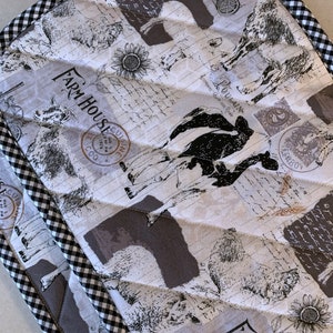 May include: A white quilted pot holder with a black and white checkered border. The pot holder features a farmhouse theme with images of cows, chickens, and a sunflower. The text "Farm House" is printed on the pot holder.