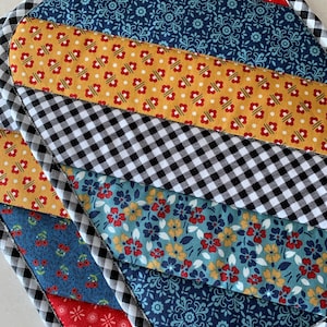 Puede incluir: Un salvamanteles acolchado con un diseño de patchwork. Presenta rayas diagonales rojas, azul marino, doradas y con estampados florales, bordeadas por cuadros vichy en blanco y negro. Los bordes están acabados con un bies en blanco y negro.