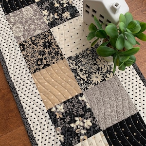 May include: A quilted table runner with a patchwork design in black, white, and beige. The runner features various floral and dotted patterns. A small white ceramic house and a potted green plant are placed on the runner.