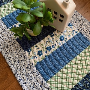 May include: A quilted table runner with a patchwork design in shades of blue, green, and white. A small white ceramic house and a potted succulent plant sit on the runner, adding a touch of decor.