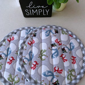 May include: Two round, quilted pot holders with a snowman pattern on a gray background. Each snowman has a black top hat, scarf, and buttons. The edges are trimmed with a gray and white checkered fabric.