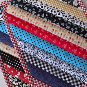 May include: A quilted, heart-shaped hot pad with a red and white gingham border. The center features diagonal strips of various patterned fabrics, including floral, polka dot, and gingham designs. The color palette includes red, blue, black, white, and yellow.
