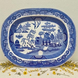 May include: An oval ceramic serving platter featuring a blue and white willow pattern. The design depicts a landscape with pagodas, a bridge, willow trees, and two birds in flight. The wide border displays a geometric and floral pattern.