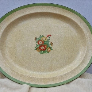 Puede incluir: Una bandeja de servicio ovalada con una base de color crema y textura finamente moteada. El borde está delimitado por una banda verde sólida. Un pequeño arreglo floral central muestra flores rojas, amarillas y verdes con hojas verdes.