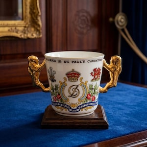 Könnte beinhalten: Ein weißer Porzellanbecher mit zwei goldfarbenen Henkeln, verziert mit einer königlichen Krone, einem roten Drachen und den verschlungenen Initialen D und C. Der Text lautet MARRIED IN ST. PAUL'S CATHEDRAL und PRINCE OF WALES AND LADY DIANA SPENCER.