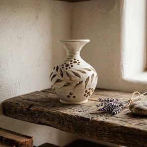May include: A cream-coloured ceramic vase with a speckled finish, featuring a narrow neck and a rounded body. The surface is decorated with cut-out leaf patterns and small circular perforations arranged in clusters around the exterior.