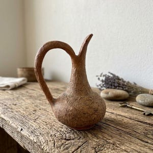 May include: A small, brown, textured pitcher with a rounded base, a slender neck, and a curved handle. The surface of the vessel has a rough, organic appearance with visible fibers and an uneven, matte finish, standing upright on a surface.