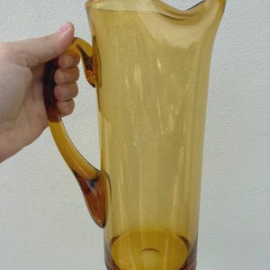 Puede incluir: Una jarra cilíndrica alta hecha de vidrio translúcido de color ámbar. El recipiente presenta un cuerpo vertical liso con un pico vertedor ancho y curvado en el borde superior y un asa resistente de vidrio ámbar a juego en el lateral.