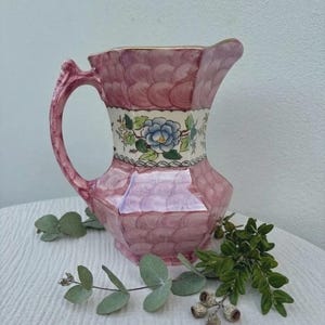 Puede incluir: Una jarra de cerámica con un patrón de ondas rosas en las secciones superior e inferior, y una banda central blanca decorada con una flor azul y hojas verdes. La jarra tiene un asa curva y un pico vertedor con un borde de tono dorado.
