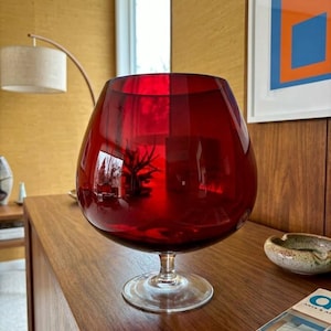 May include: A large, deep red glass vessel shaped like a brandy snifter, featuring a wide, rounded bowl that tapers toward a clear glass stem and a circular base. The smooth, translucent red glass reflects light, creating a vibrant, glossy appearance.