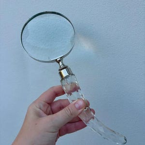 May include: A magnifying glass featuring a circular lens encased in a silver-toned metal frame, connected to a curved, faceted clear glass handle that tapers to a point, reflecting light through its geometric, multi-sided surface structure.