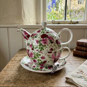 May include: A porcelain tea set for one featuring a white base with a dense pattern of pink and dark red roses and green leaves. The set includes a teapot with a lid, a matching teacup, and a saucer, all decorated with the same floral motif.