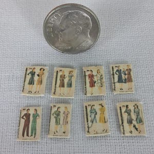 May include: Eight miniature rectangular paper cutouts featuring vintage fashion illustrations of dresses and suits, arranged in two rows below a silver United States dime coin for scale, each showing detailed garment designs and patterns.