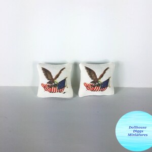 May include: Two small, square pillows with a patriotic design. Each pillow features a brown eagle holding a red, white, and blue American flag. The pillows are on a light gray surface.