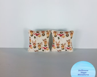 May include: Two miniature Christmas-themed pillows. The pillows feature a cream-colored background with a repeating pattern of reindeer, Santa hats, and holly. The pillows are square-shaped and have a white border.