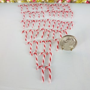 May include: A collection of miniature red and white candy canes arranged in a triangular pattern on a white surface. A US dime is included for scale.