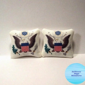 May include: Two white miniature pillows with embroidered images of the Great Seal of the United States. The seal features an eagle with outstretched wings, a shield with red and white stripes, and olive branches and arrows.