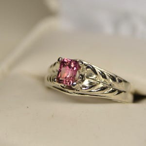 May include: A silver ring with a square, pink gemstone. The band features a detailed leaf design. The ring is set against a soft, off-white background, highlighting the jewelry's intricate details.