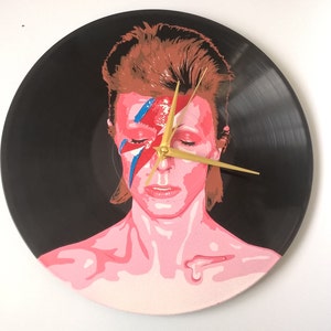 May include: A black vinyl record clock featuring a painted portrait of David Bowie with his signature lightning bolt makeup. The clock hands are gold.