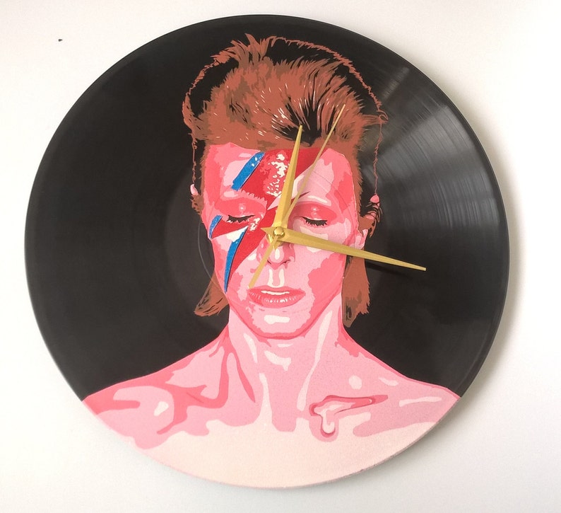 May include: A black vinyl record clock featuring a painted portrait of David Bowie with his signature lightning bolt makeup. The clock hands are gold.