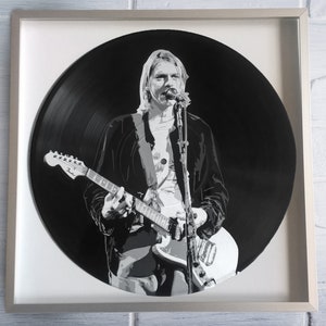 Puede incluir: Un disco de vinilo en blanco y negro con un retrato de un músico tocando una guitarra. El músico lleva una chaqueta negra y una camiseta blanca.