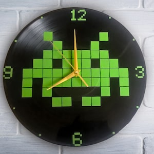 Puede incluir: Un reloj de vinilo negro con un diseño de invasor espacial pixelado verde y agujas doradas. La esfera del reloj tiene los números 12, 3, 6 y 9.