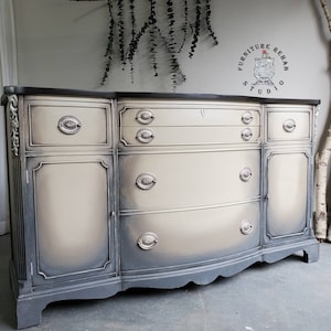 May include: A vintage-style buffet with a curved front and a distressed gray and white paint finish. The buffet has four drawers and two cabinet doors with ornate hardware. The top of the buffet is black.