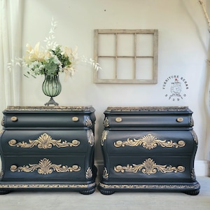 May include: Two matching dark charcoal nightstands with two drawers each, featuring ornate gold-painted scrollwork carvings on the drawer fronts and edges. Each piece has a brown marble-patterned top and small round gold knobs on the top drawer.