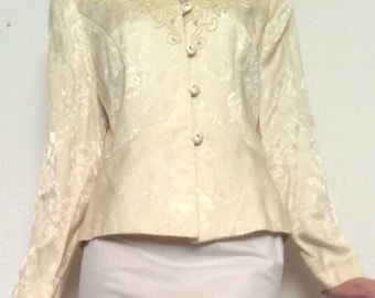 May include: A cream-colored, long-sleeved jacket with a floral pattern and pearl-like embellishments. The jacket has a button-up front and a fitted silhouette. The model is also wearing a white slip skirt with lace trim.