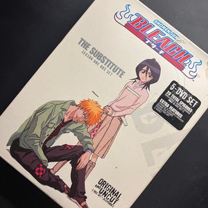Könnte beinhalten: Ein weißes DVD-Box-Set für die Anime-Serie Bleach. Die Box zeigt den Titel "Bleach" in rot-weißen Buchstaben und den Untertitel "The Substitute" in schwarzen Buchstaben. Die Box zeigt auch Bilder der Charaktere Ichigo Kurosaki und Rukia Kuchiki. Das Box-Set enthält 5 DVDs und 20 Episoden, einschließlich Bonus-Episoden.