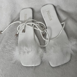 May include: A pair of white sandals with a square toe design. The sandals feature a strap that ties around the ankle and are adorned with white feathers. The word "FASHION" is printed on the footbed.