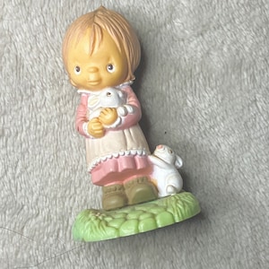 May include: A small figurine of a girl in a pink dress holding a white bunny. She is standing on a green base with a white bunny beside her.