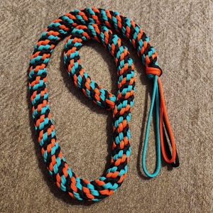 May include: Braided rope leash in a vibrant pattern of black, orange, and turquoise. The leash is looped and has a loop at the end with turquoise and orange strands. The rope is lying on a textured, brown surface.