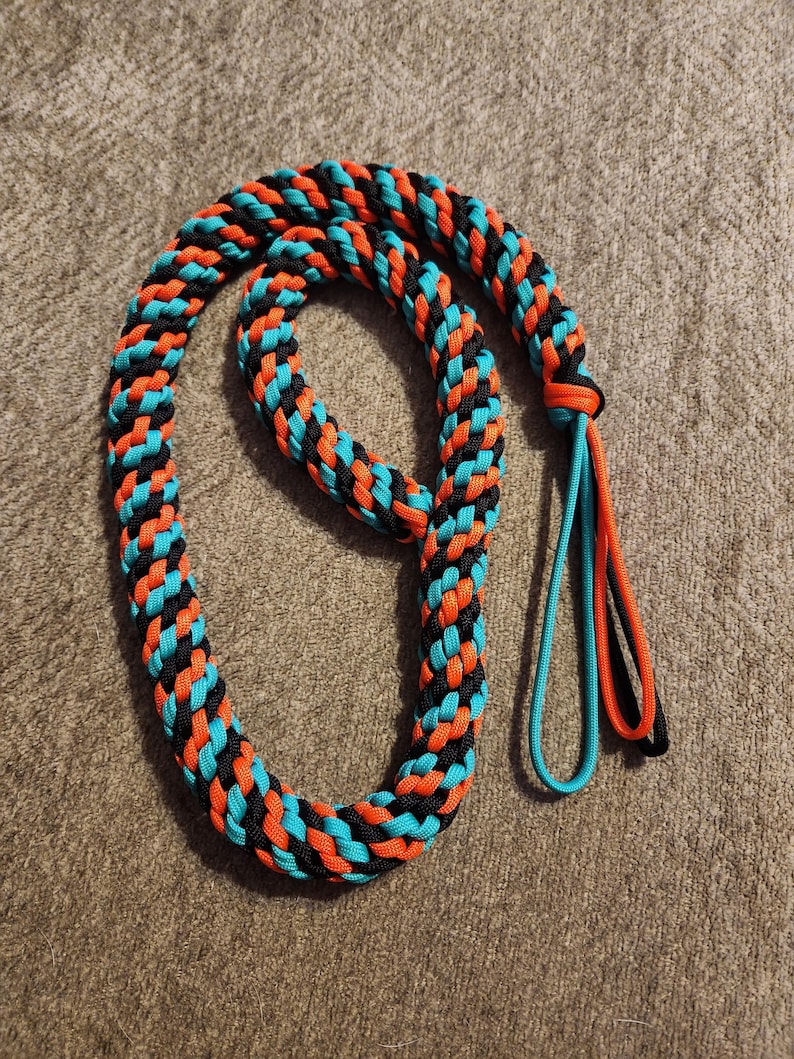May include: Braided rope leash in a vibrant pattern of black, orange, and turquoise. The leash is looped and has a loop at the end with turquoise and orange strands. The rope is lying on a textured, brown surface.
