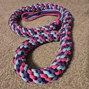 May include: A figure-eight shaped braided rope, crafted from pink, purple, and light blue cords. The rope is tightly woven, creating a textured surface. The ends are tied together with a loop. Likely a pet accessory.