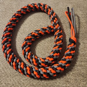 May include: A coiled rope made from interwoven orange, black, and grey strands. The rope has a loop at one end and a tassel of three cords at the other. The background is a neutral, textured surface.