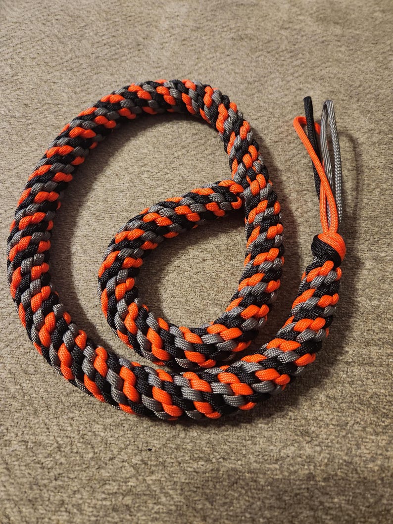 May include: A coiled rope made from interwoven orange, black, and grey strands. The rope has a loop at one end and a tassel of three cords at the other. The background is a neutral, textured surface.