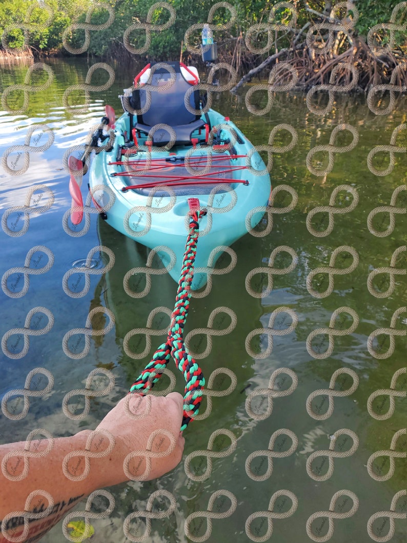 May include: A light blue kayak with red detailing and a braided rope. The kayak is in the water near mangroves. A hand holds the black, red, and green braided rope.