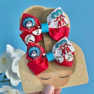 May include: Two hair bows featuring a red and white ribbon pattern with blue-haired characters and white circles containing the text Thing 1 and Thing 2. The bows are secured with a bright blue centre band and mounted on a tan display card.