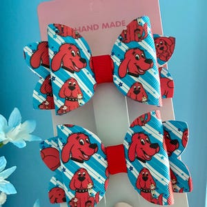 May include: Two hair bows attached to a pink display card labelled HAND MADE. Each bow features a repeating pattern of a red dog character on a light blue background with white diagonal stripes and small blue stars, centred with a red ribbon.