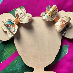May include: Two hair bows featuring a cream-coloured pattern of Bambi and Thumper characters, accented with a solid brown centre band. The bows are attached to a light-coloured wooden display cutout shaped like a head with two round ears.