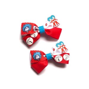 May include: A pair of red hair bows with blue centres. Each bow features a Dr. Seuss character design, with the words "Thing 1" and "Thing 2" printed on them. These bows are likely for children and are set against a white background.