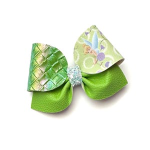 May include: A green hair bow with a layered design. The bow features a woven texture on one side and a Tinkerbell print on the other. A sparkly, light blue accent is in the centre. The bow is likely for a child.